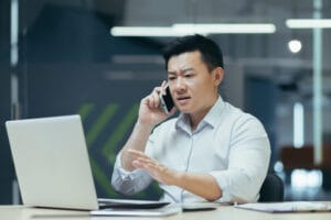 Worried and nervous young Asian man talking on the phone in the office, looking at the laptop, solving problems, questions